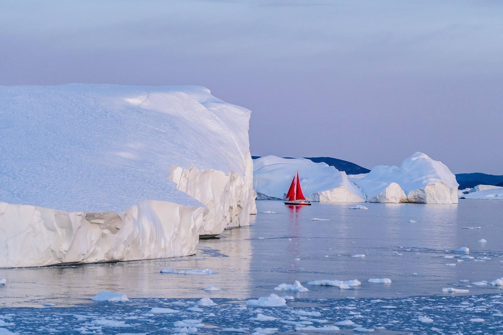 A sailboat glides past towering icebergs in Greenland.