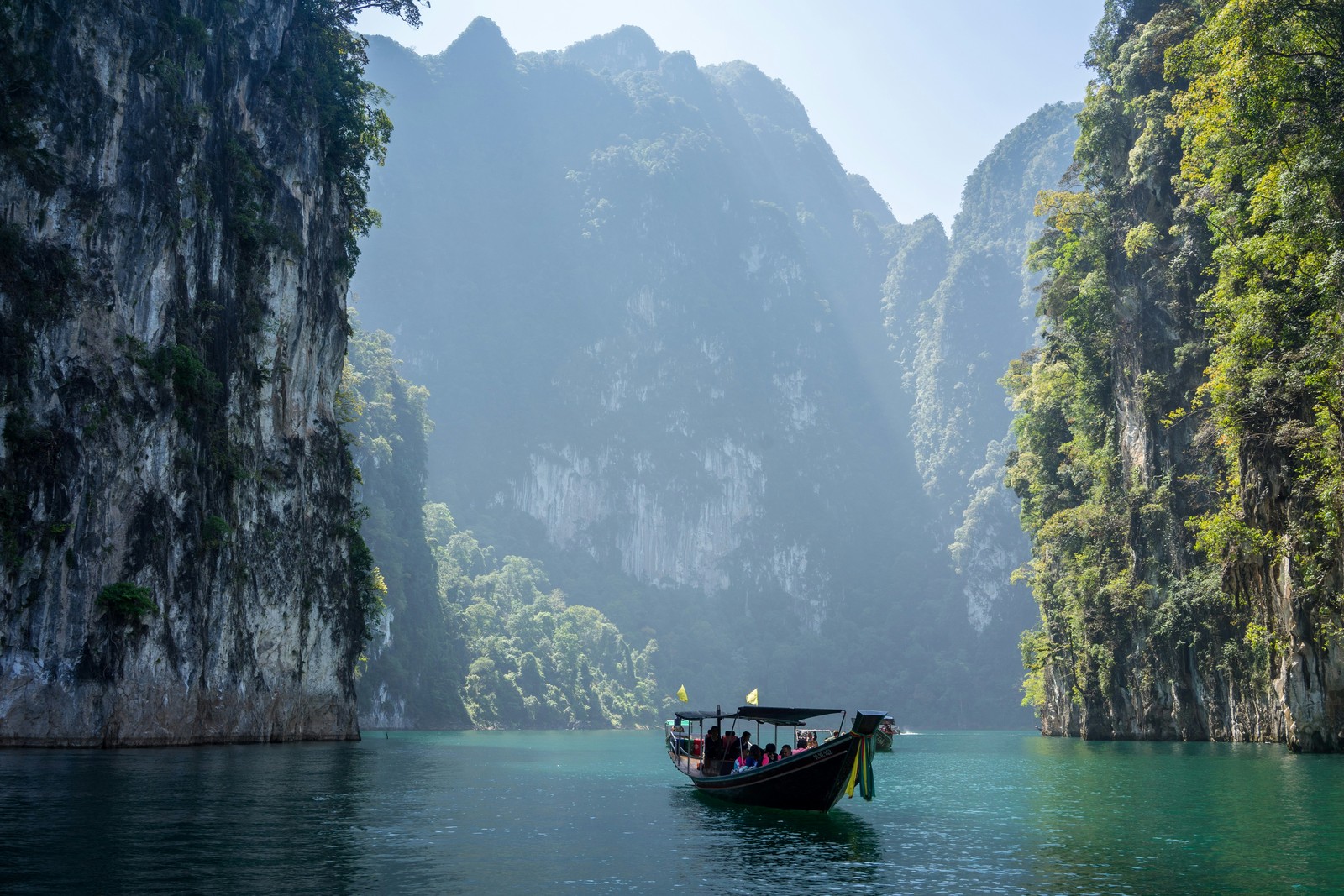 A long-tail boat glides through emerald waters between towering limestone cliffs in Khao Sok National Park. Photo by Robin Noguier.